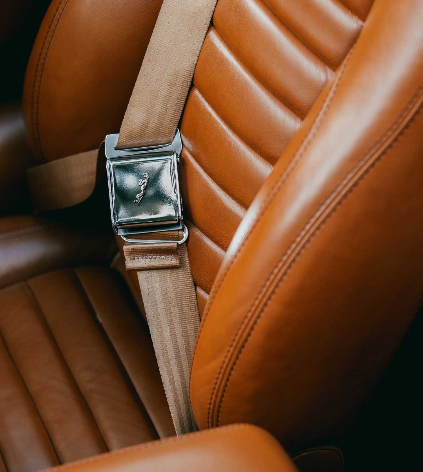 A closeup of a tanned leather seat with a focus on the seatbelt buckle with the jaguar emblem extruded on the metalwork